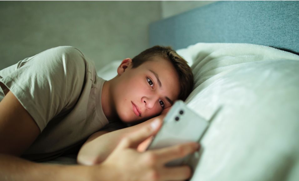 Adolescent ayant l'âge de la majorité numérique dans son lit avec un smartphone à la main, scrollant les réseaux sociaux. 